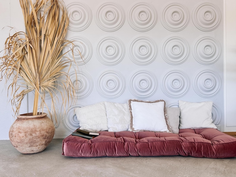 May include: A living room scene featuring a low, burgundy velvet sofa with several white and beige pillows. A large, dried palm frond is in a terracotta pot. The wall has a repeating circular pattern.