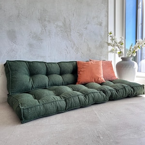 Può includere: Un divano da pavimento in tessuto verde scuro strutturato. Il divano ha un design trapuntato ed è abbinato a due cuscini decorativi color pesca. L'ambiente è una stanza luminosa con una parete di colore neutro e un vaso di fiori bianchi.