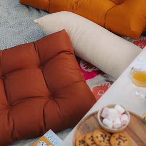 Peut inclure: Sc&egrave;ne de pique-nique en ext&eacute;rieur avec coussins orange et rouille, un traversin cr&egrave;me et une table blanche. Un plateau en bois contient des biscuits et des guimauves. Un jeu de Yahtzee est visible.