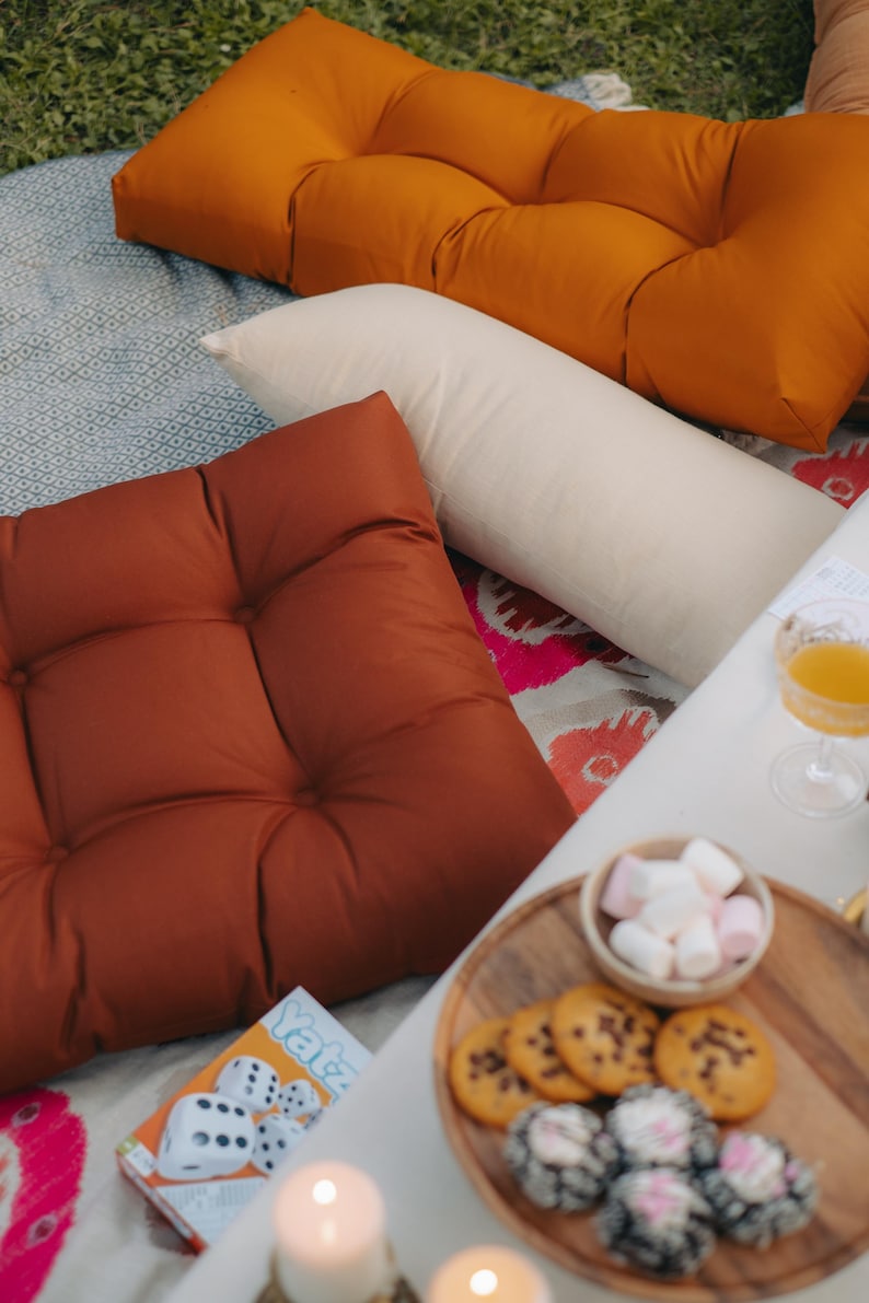 Peut inclure: Sc&egrave;ne de pique-nique en ext&eacute;rieur avec coussins orange et rouille, un traversin cr&egrave;me et une table blanche. Un plateau en bois contient des biscuits et des guimauves. Un jeu de Yahtzee est visible.