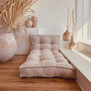 May include: A light pink tufted floor cushion with a rectangular shape. The cushion is placed on a wooden floor. Decorative vases and dried plants are in the background, creating a neutral, bohemian aesthetic.