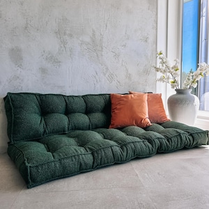 May include: A dark green tufted floor sofa with two orange throw pillows. The sofa is rectangular and has a textured, plush appearance. A white vase with white flowers sits near a window.