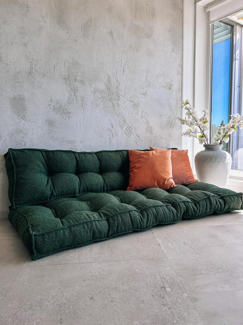 May include: A dark green tufted floor sofa with two orange throw pillows. The sofa is rectangular and has a textured, plush appearance. A white vase with white flowers sits near a window.
