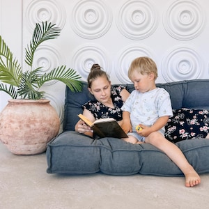 May include: Two people are seated on a gray floor cushion, reading a book. The person on the left is wearing a black floral top, and the person on the right is wearing a light blue t-shirt. A large terracotta pot with a green plant is to the left.