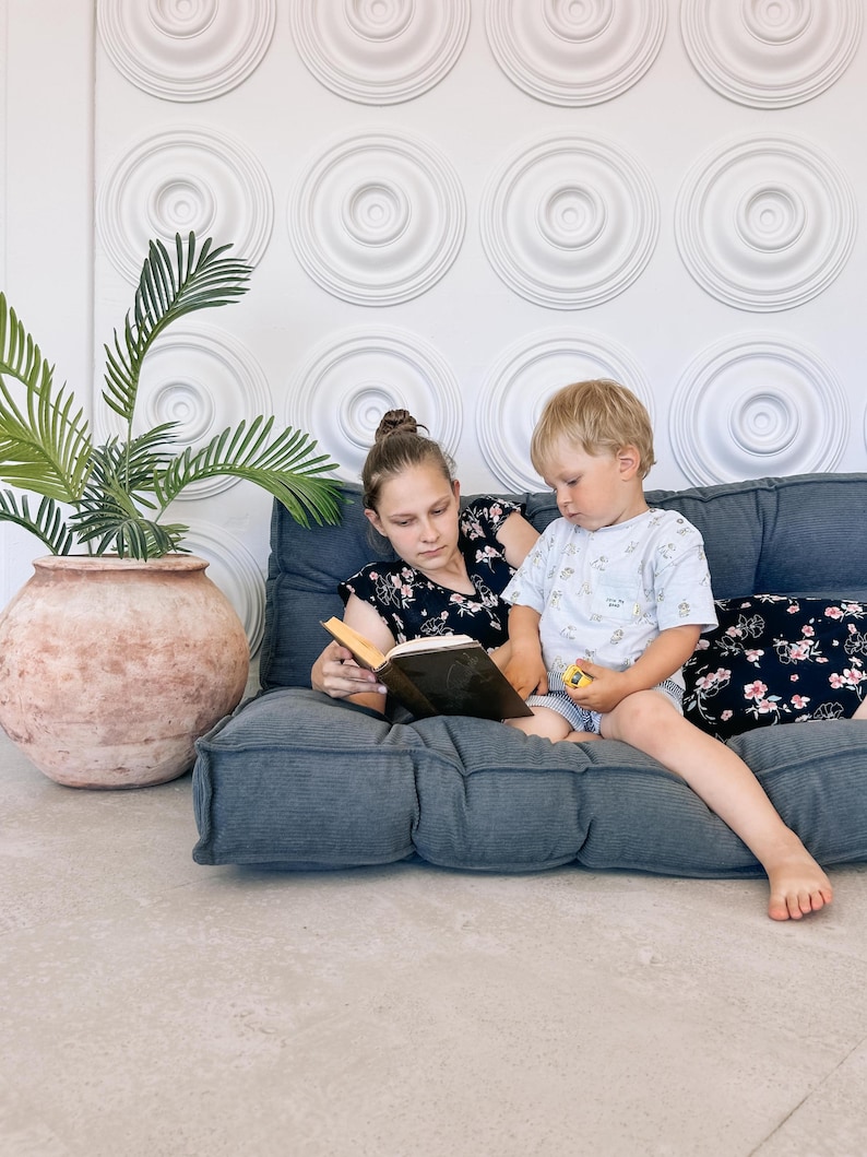 May include: Two people are seated on a gray floor cushion, reading a book. The person on the left is wearing a black floral top, and the person on the right is wearing a light blue t-shirt. A large terracotta pot with a green plant is to the left.