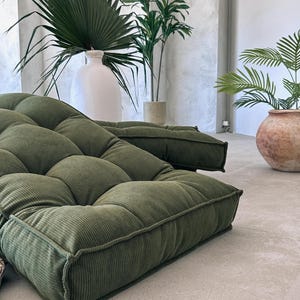May include: Two olive green corduroy floor cushions with a tufted design. The cushions are rectangular and have a thick, textured appearance. The background includes large potted plants and a white vase.