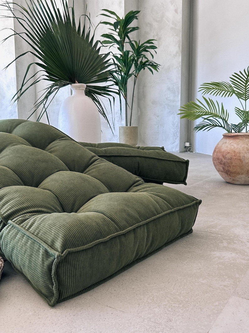 May include: Two olive green corduroy floor cushions with a tufted design. The cushions are rectangular and have a thick, textured appearance. The background includes large potted plants and a white vase.
