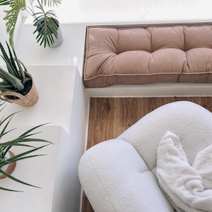May include: An overhead shot of a cozy interior space. A white, rounded chair with a white blanket sits on a wooden floor. A pink, tufted cushion is against a white wall. Potted plants add a touch of greenery.