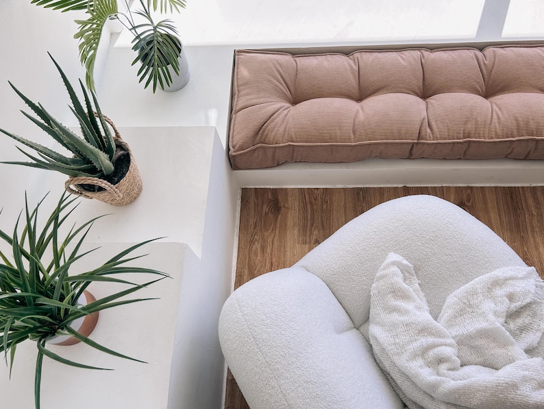 May include: An overhead shot of a cozy interior space. A white, rounded chair with a white blanket sits on a wooden floor. A pink, tufted cushion is against a white wall. Potted plants add a touch of greenery.