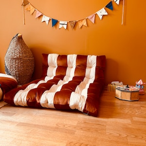 Peut inclure: Un coussin de sol rayé brun et blanc avec une texture en velours. Le coussin est placé sur un sol en bois dans une pièce avec un mur orange clair. Une guirlande de drapeaux colorés est accrochée au mur au-dessus du coussin.