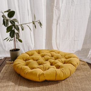 Peut inclure: Un coussin de sol rond jaune moutarde avec un design capitonn&eacute; est pos&eacute; sur un tapis tiss&eacute;. Une plante en pot avec des feuilles vertes est &agrave; gauche, avec des rideaux blancs en arri&egrave;re-plan.