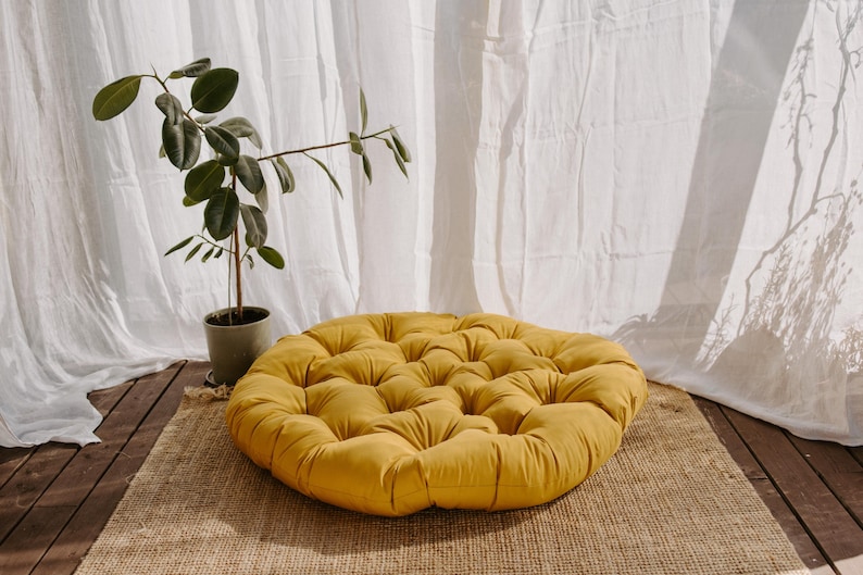 Peut inclure: Un coussin de sol rond jaune moutarde avec un design capitonn&eacute; est pos&eacute; sur un tapis tiss&eacute;. Une plante en pot avec des feuilles vertes est &agrave; gauche, avec des rideaux blancs en arri&egrave;re-plan.