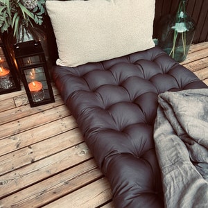 Dark gray tufted outdoor mattress cushion styled as a single bed setup, paired with a cream textured pillow and soft gray blanket. Large Scandinavian patio cushion made from durable water-repellent fabric, suitable for terraces, balconies and garden lounges. Designed for comfortable outdoor relaxation while maintaining a clean, modern Nordic aesthetic.