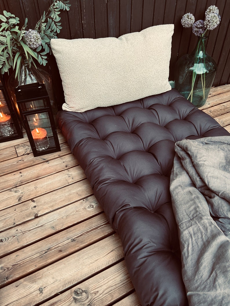 Dark gray tufted outdoor mattress cushion styled as a single bed setup, paired with a cream textured pillow and soft gray blanket. Large Scandinavian patio cushion made from durable water-repellent fabric, suitable for terraces, balconies and garden lounges. Designed for comfortable outdoor relaxation while maintaining a clean, modern Nordic aesthetic.