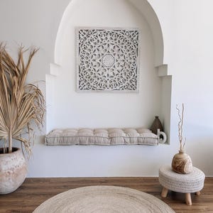 May include: A neutral-toned interior scene featuring a square, white, carved wooden wall panel with a floral design. A beige cushion sits below the panel. A round, woven rug and a small, wooden stool with a woven top are also present.