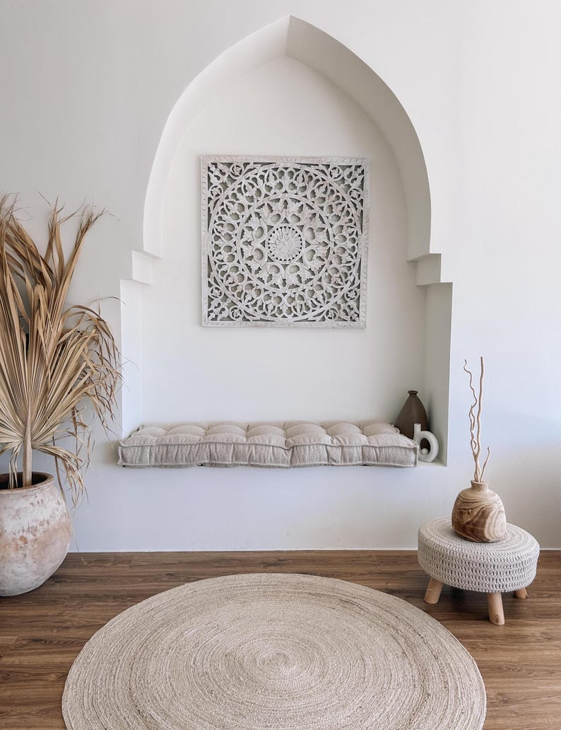 May include: A neutral-toned interior scene featuring a square, white, carved wooden wall panel with a floral design. A beige cushion sits below the panel. A round, woven rug and a small, wooden stool with a woven top are also present.