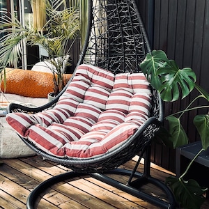 Puede incluir: Una silla colgante negra con un coj&iacute;n a rayas rojas y blancas. La silla est&aacute; en una terraza de madera con una planta verde en el fondo.