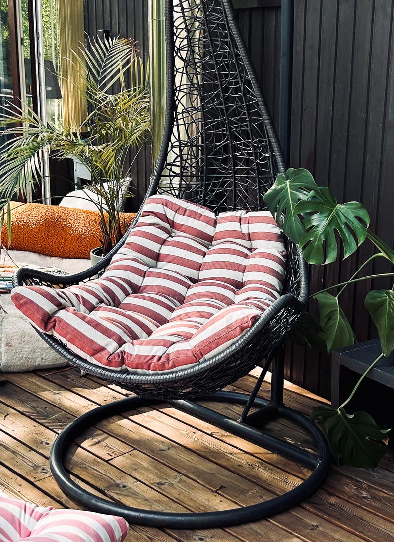 Puede incluir: Una silla colgante negra con un coj&iacute;n a rayas rojas y blancas. La silla est&aacute; en una terraza de madera con una planta verde en el fondo.