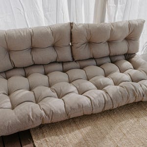 A beige tufted outdoor cushion set. The set includes a backrest and a seat cushion, both with a grid pattern. The cushions are made of a textured fabric and are placed on a woven rug. The background features white curtains and natural light.