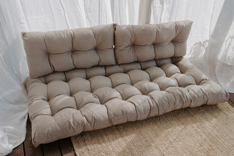 A beige tufted outdoor cushion set. The set includes a backrest and a seat cushion, both with a grid pattern. The cushions are made of a textured fabric and are placed on a woven rug. The background features white curtains and natural light.