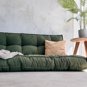 May include: A low-profile, olive green corduroy sofa with a tufted design. A tan pillow rests on the sofa, and a patterned throw is draped over the side. A potted plant sits on a small wooden side table.