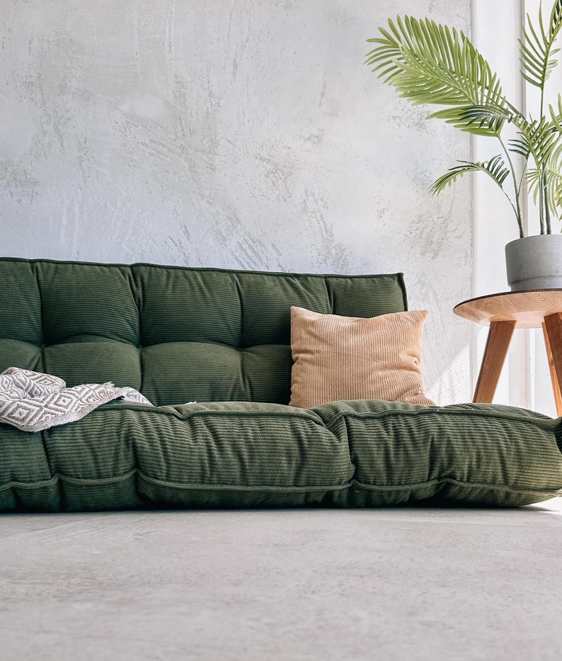 May include: A low-profile, olive green corduroy sofa with a tufted design. A tan pillow rests on the sofa, and a patterned throw is draped over the side. A potted plant sits on a small wooden side table.