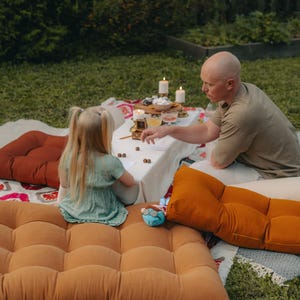 Outdoor scene with a picnic setup. A person and a child are seated on large, tufted floor cushions in shades of orange and brown. A white tablecloth is set with food and candles. A blue stuffed animal is visible.