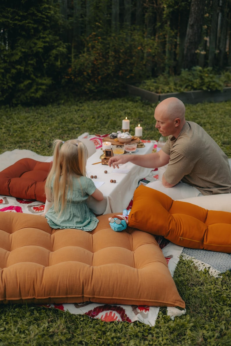 Outdoor scene with a picnic setup. A person and a child are seated on large, tufted floor cushions in shades of orange and brown. A white tablecloth is set with food and candles. A blue stuffed animal is visible.