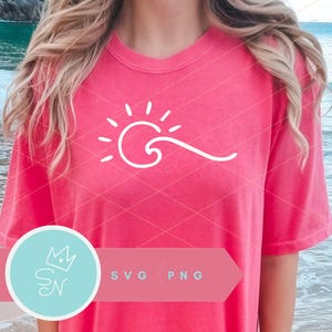 Puede incluir: Una mujer lleva una camiseta rosa con un diseño de sol y ola blanco. El diseño es simple y minimalista. La camiseta es una gran opción para un día de verano en la playa.