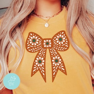 Op de afbeelding: Mosterdgeel T-shirt met een groot, sierlijk strikdesign in bruin, wit en groen. De strik heeft een bloemenpatroon. Het T-shirt is een casual stijl.