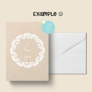 Puede incluir: Una tarjeta de felicitaci&oacute;n beige con una corona floral blanca y el texto "I love you". Una sobre blanco est&aacute; al lado de la tarjeta. La palabra "Example" est&aacute; escrita encima de la tarjeta.