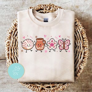 May include: A beige crewneck sweatshirt featuring a graphic print of four anthropomorphic characters: a concha bread, a clay jar, a flower, and a butterfly. Each character has a smiling face, arms, and legs, surrounded by small pink hearts and flowers.