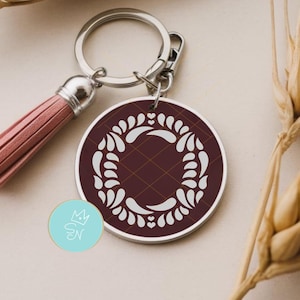 May include: A round burgundy keychain with a silver border and a white floral design. The keychain has a pink tassel and a silver keyring. The background includes wheat stalks.