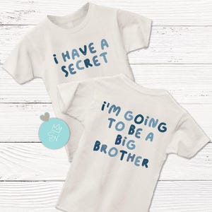 May include: Two white t-shirts with blue text. One shirt says "I HAVE A SECRET" and the other says "I'M GOING TO BE A BIG BROTHER". The shirts are likely for a pregnancy announcement or a sibling reveal.