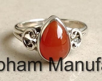 May include: A silver ring with a teardrop-shaped, orange-red gemstone. The ring has decorative scrollwork on either side of the gemstone and the text "Shubham Manufacturers" below the ring.