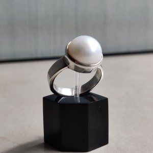 May include: A silver ring with a large white pearl set in a bezel setting. The ring is on a black display stand.
