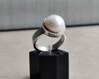 May include: A silver ring with a large white pearl set in a bezel setting. The ring is on a black display stand.