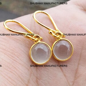 May include: A pair of gold-colored drop earrings with round, light gray gemstones. The earrings have a simple design with a hook closure. The text "SHUBHAM MANUFACTURERS" is visible on the image.