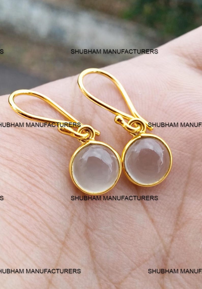 May include: A pair of gold-colored drop earrings with round, light gray gemstones. The earrings have a simple design with a hook closure. The text "SHUBHAM MANUFACTURERS" is visible on the image.