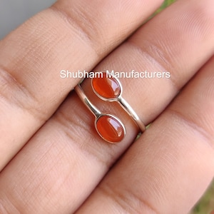 Può includere: Un anello d'argento con due pietre di corniola arancione. L'anello è di stile avvolgente e viene mostrato su un dito.