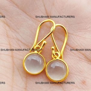 May include: A pair of gold-colored drop earrings with a round, light gray stone. The earrings have a simple, elegant design with a hook closure. The text "SHUBHAM MANUFACTURERS" is visible in the background.