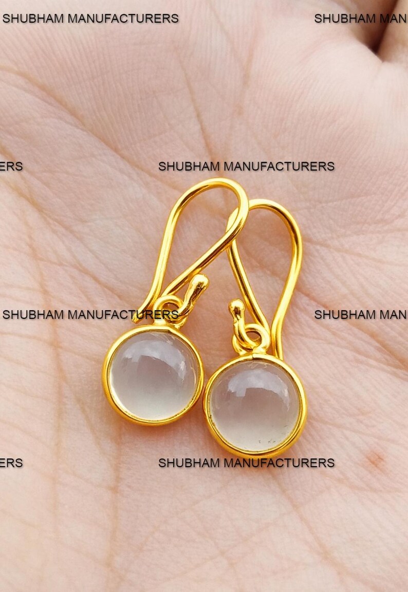 May include: A pair of gold-colored drop earrings with a round, light gray stone. The earrings have a simple, elegant design with a hook closure. The text "SHUBHAM MANUFACTURERS" is visible in the background.