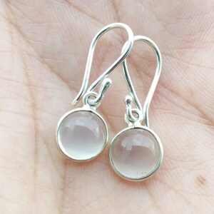 May include: A pair of silver-toned drop earrings. Each earring features a round, pale pink gemstone set in a silver bezel. The earrings have a simple, elegant design with a hook closure.