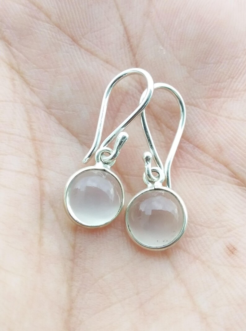 May include: A pair of silver-toned drop earrings. Each earring features a round, pale pink gemstone set in a silver bezel. The earrings have a simple, elegant design with a hook closure.