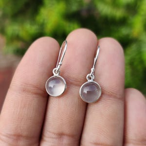 May include: A pair of silver drop earrings with round, translucent, pale purple gemstones. The earrings are held up by a person's hand, with a blurred green background.