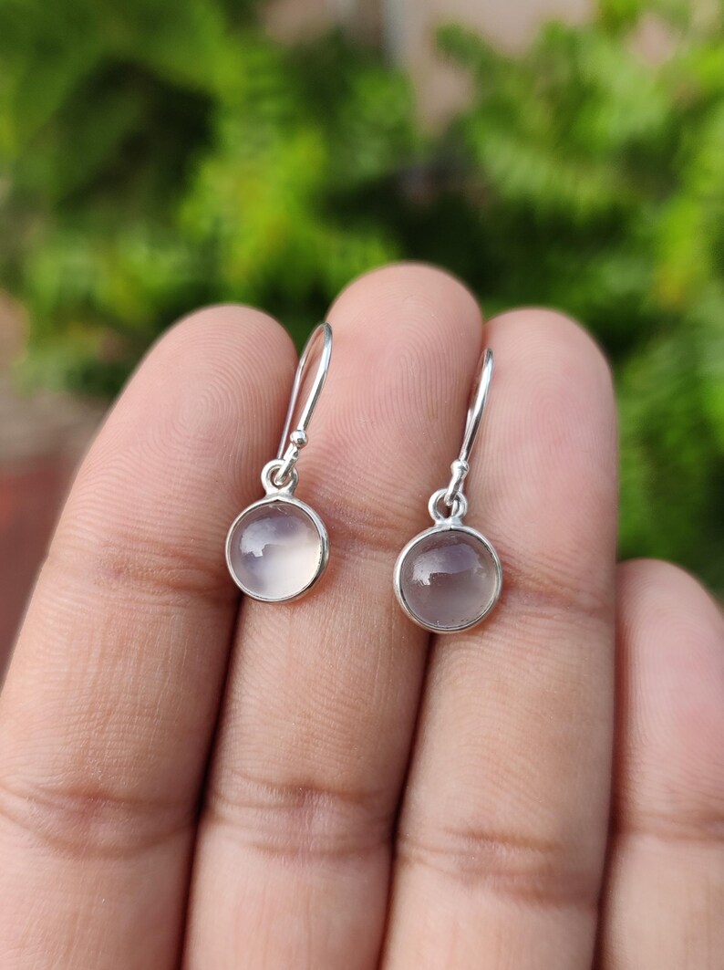 May include: A pair of silver drop earrings with round, translucent, pale purple gemstones. The earrings are held up by a person's hand, with a blurred green background.