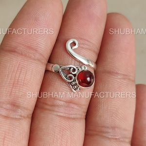 May include: A silver ring with a red gemstone. The ring features an open design with a swirling accent and decorative filigree around the gemstone. The gemstone is a deep red color and is round in shape.