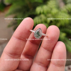 May include: A silver ring featuring a teardrop-shaped moss agate gemstone with green inclusions. The metal band is thin and polished. The text SHUBHAM MANUFACTURERS is repeated multiple times across the image. The stone has a smooth surface.