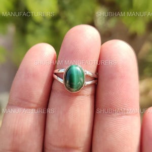 Puede incluir: Un anillo de plata con una piedra de malaquita verde de forma ovalada. El anillo tiene un diseño de banda dividida, con la piedra engastada en un bisel. La malaquita muestra un patrón de bandas. El anillo se muestra en un dedo.
