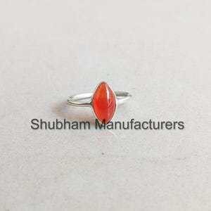 Puede incluir: Un anillo de plata con una piedra preciosa de cornalina roja. El anillo está engastado en un diseño simple y elegante.
